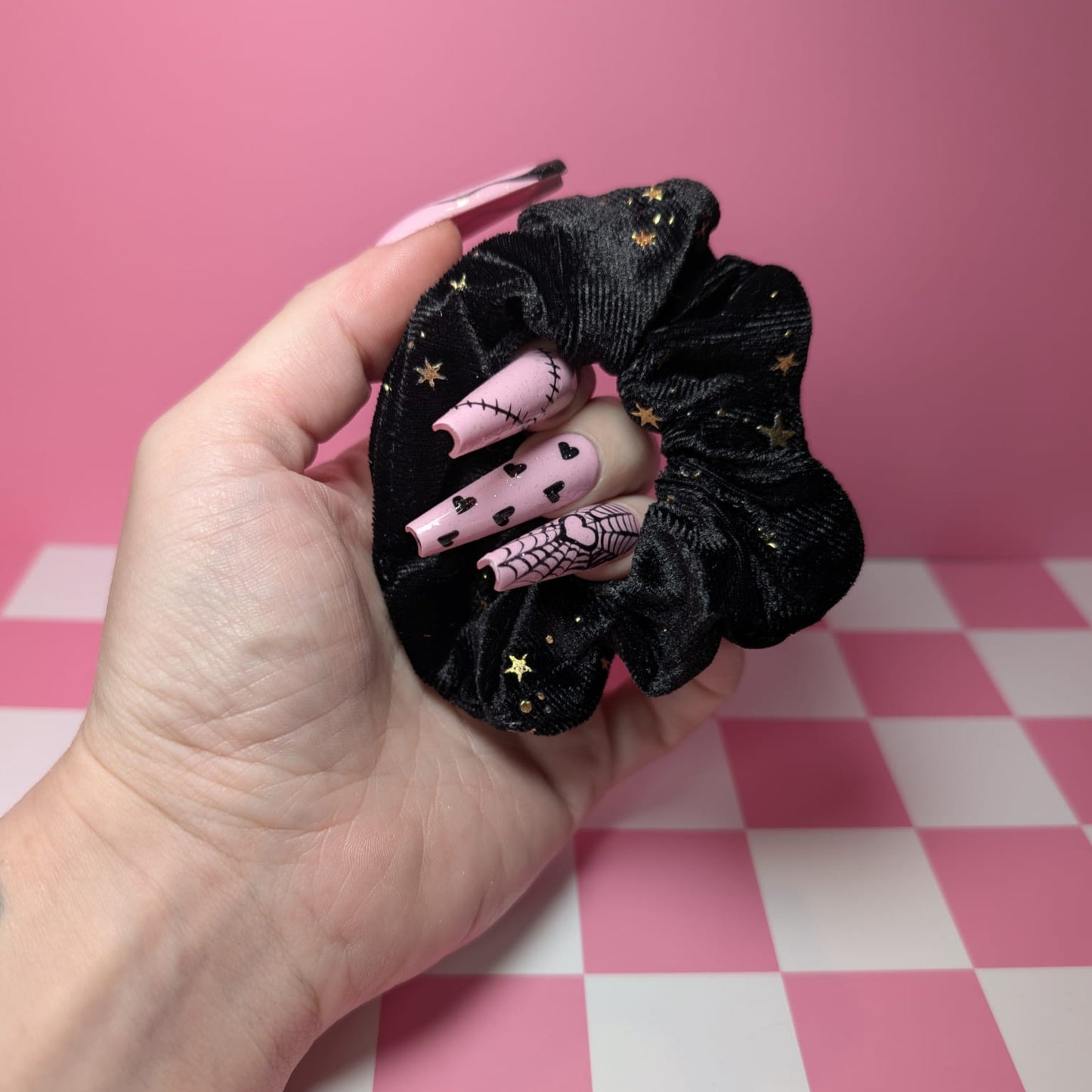 Hand holding a black scrunchie with pink and gold design against a pink and white checkered background