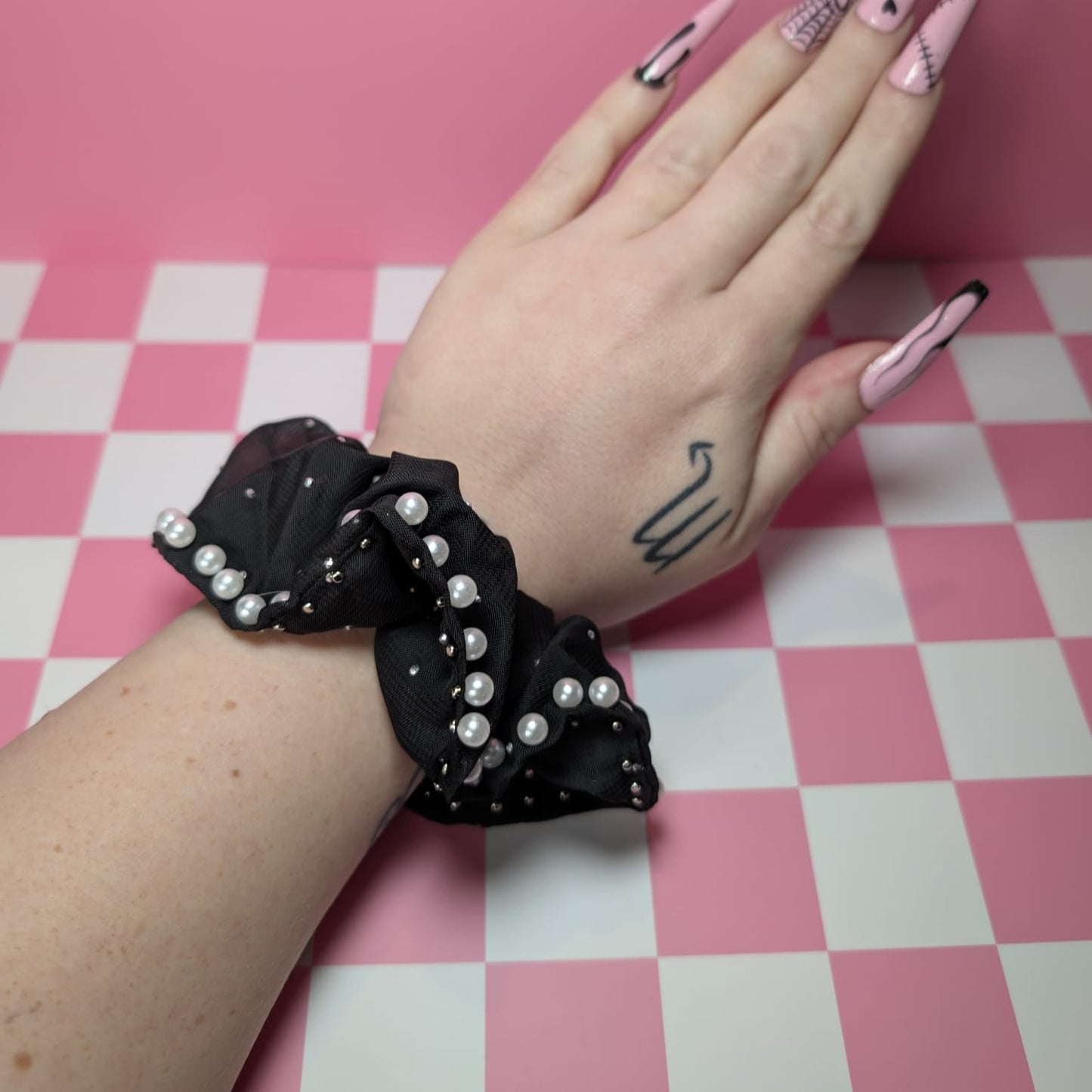 Hand wearing a black scrunchie with pearls on a pink and white checkered background