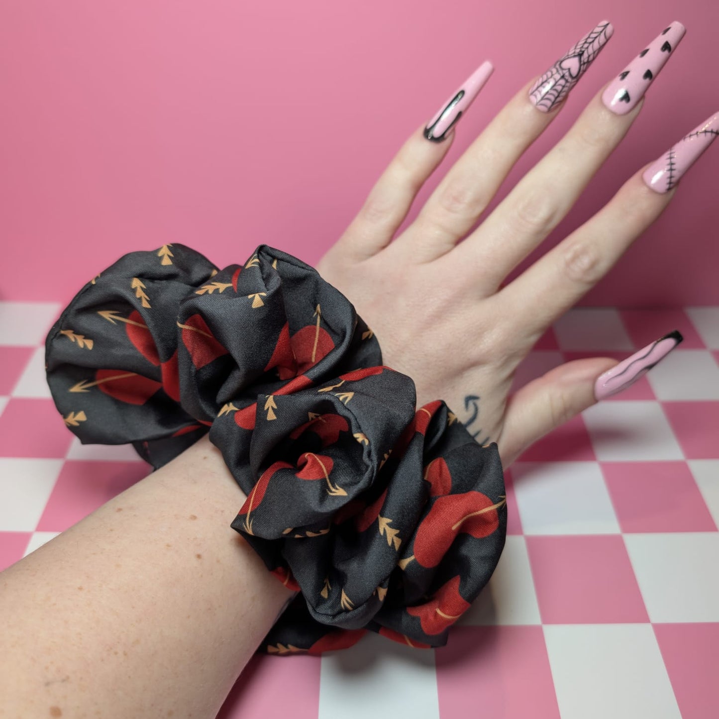 Hand wearing a black scrunchie with red patterns on a pink and white checkered background