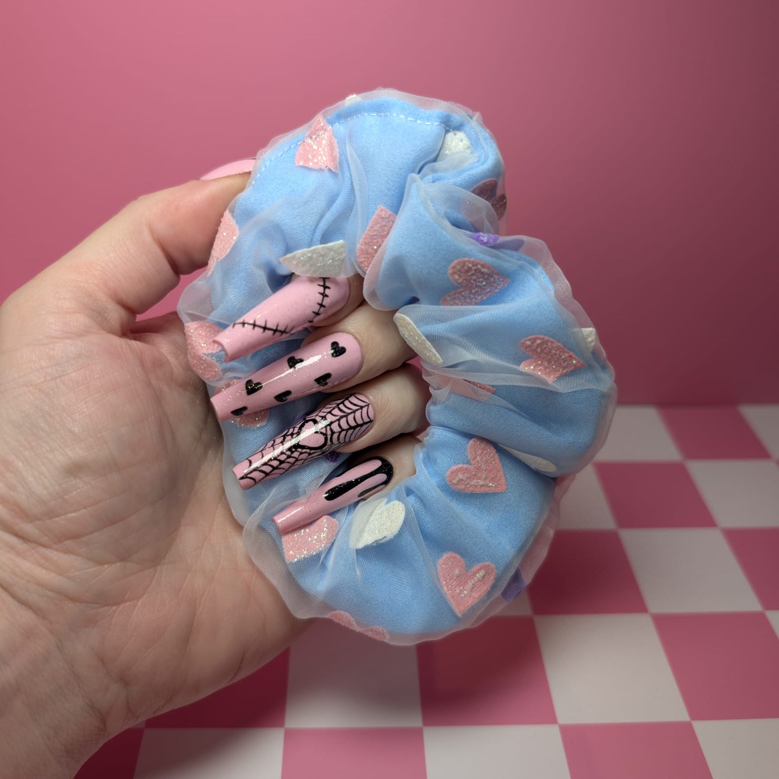Hand holding a blue scrunchie with cartoon character designs on a pink and white checkered background.