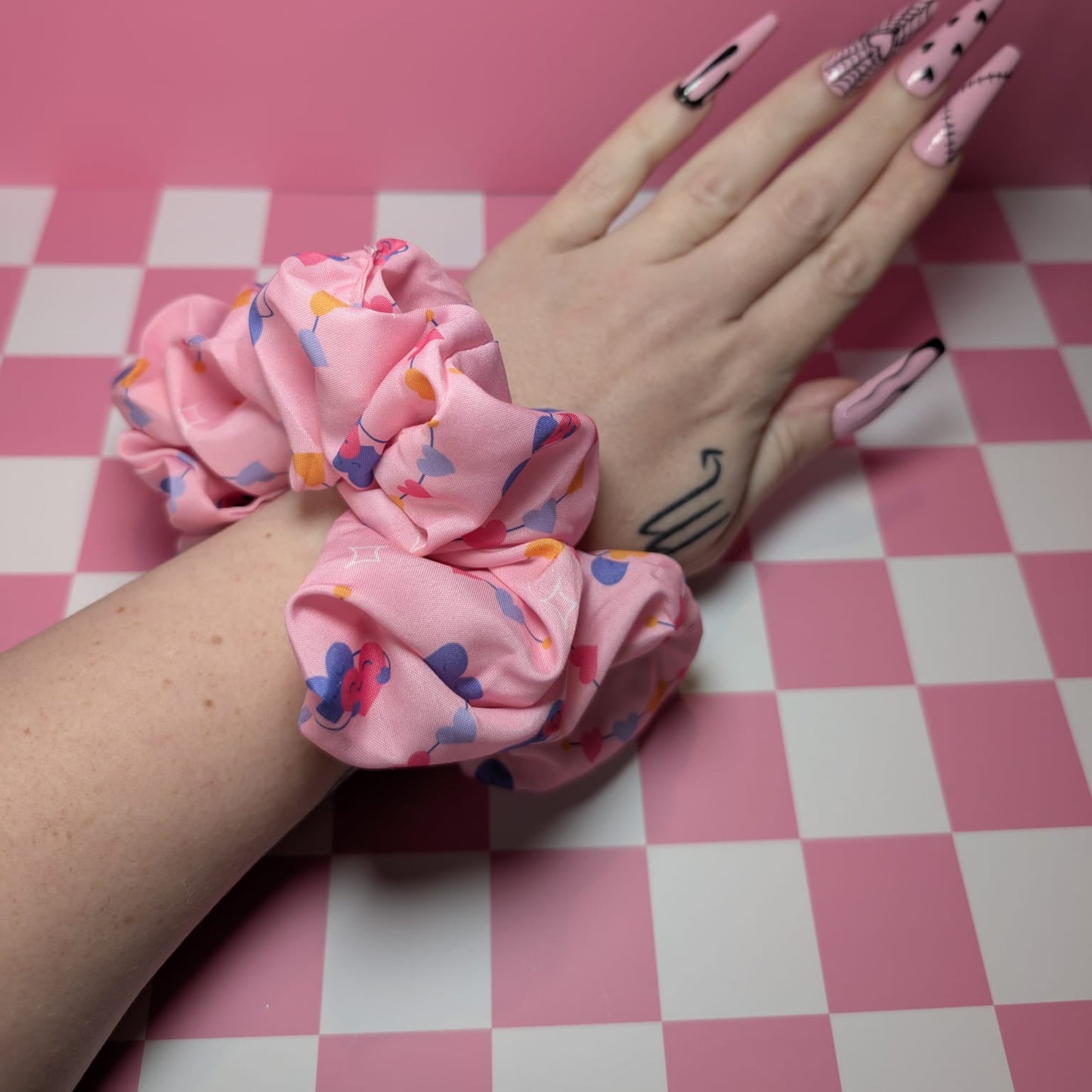 Hand wearing a pink scrunchie with colorful patterns on a pink and white checkered background
