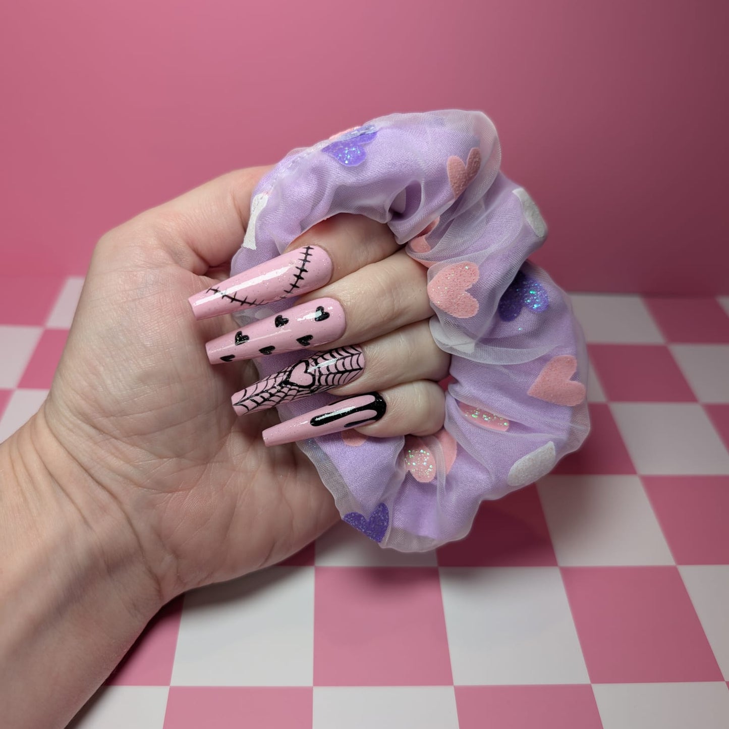Hand holding a purple scrunchie with heart patterns on a pink and white checkered surface.