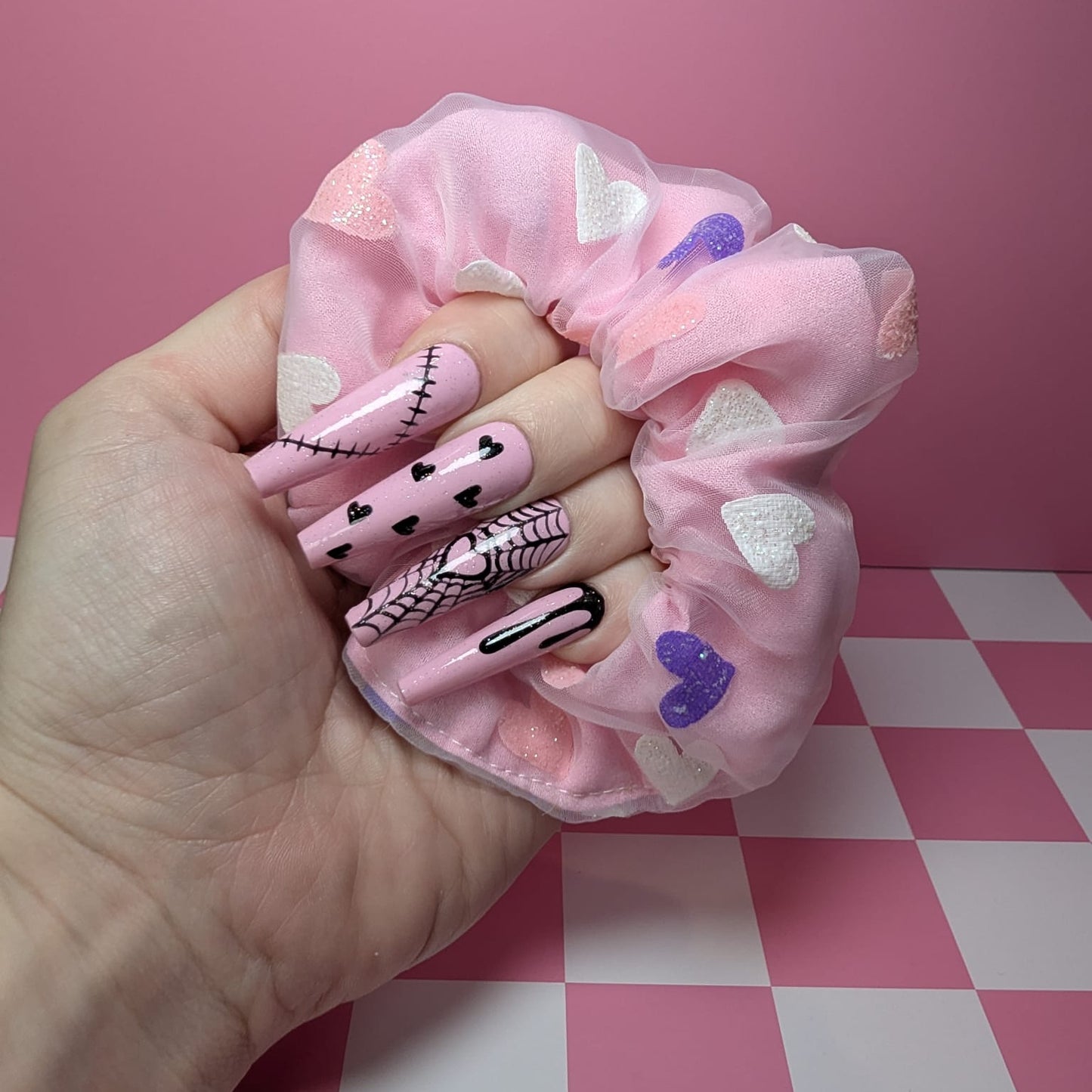 Hand holding a pink scrunchie with decorative nails on a pink and white checkered surface.