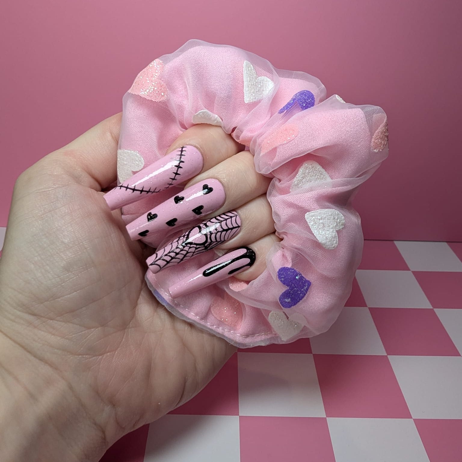 Hand holding a pink scrunchie with decorative nails on a pink and white checkered surface.