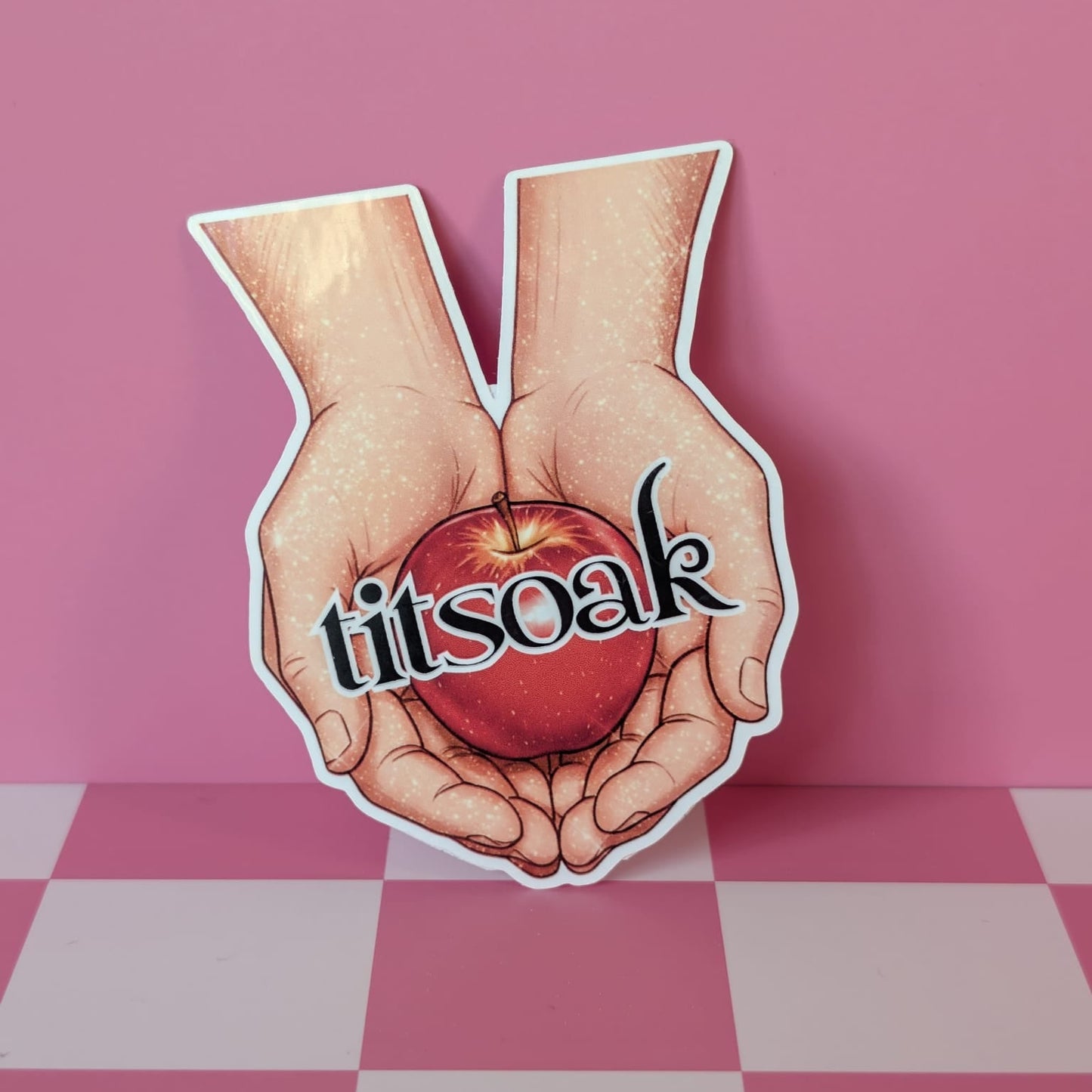 Sticker of hands holding an apple with 'titsoak' text on a pink and white checkered background