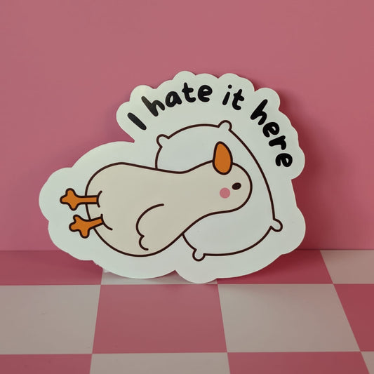 Sticker with a cartoon character and 'I hate it here' text on a pink and checkered surface
