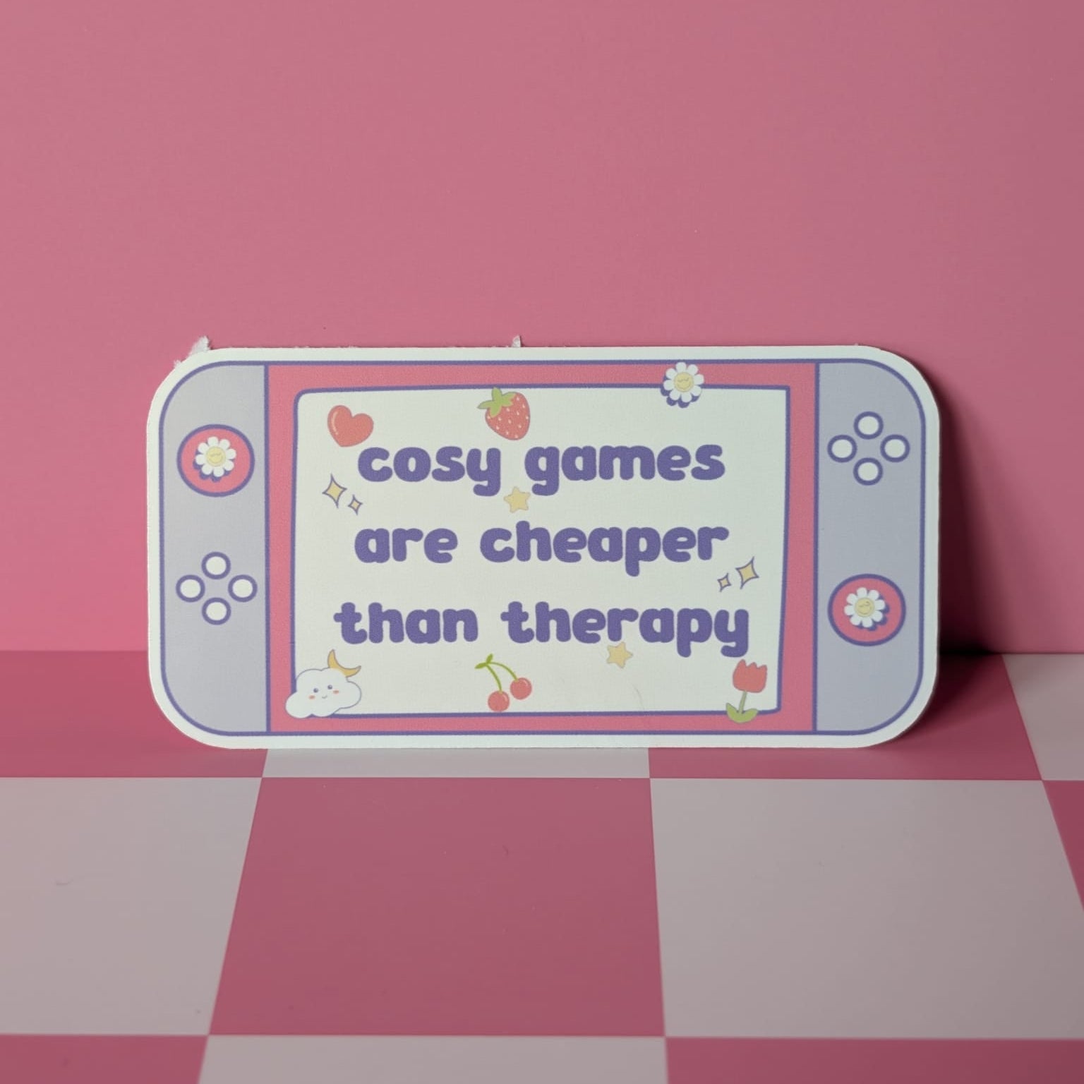 Gaming console-shaped sign with 'cosy games are cheaper than therapy' text on a pink checkered floor.
