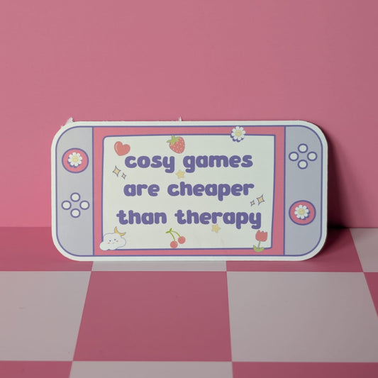 Gaming console-shaped sign with 'cosy games are cheaper than therapy' text on a pink checkered floor.